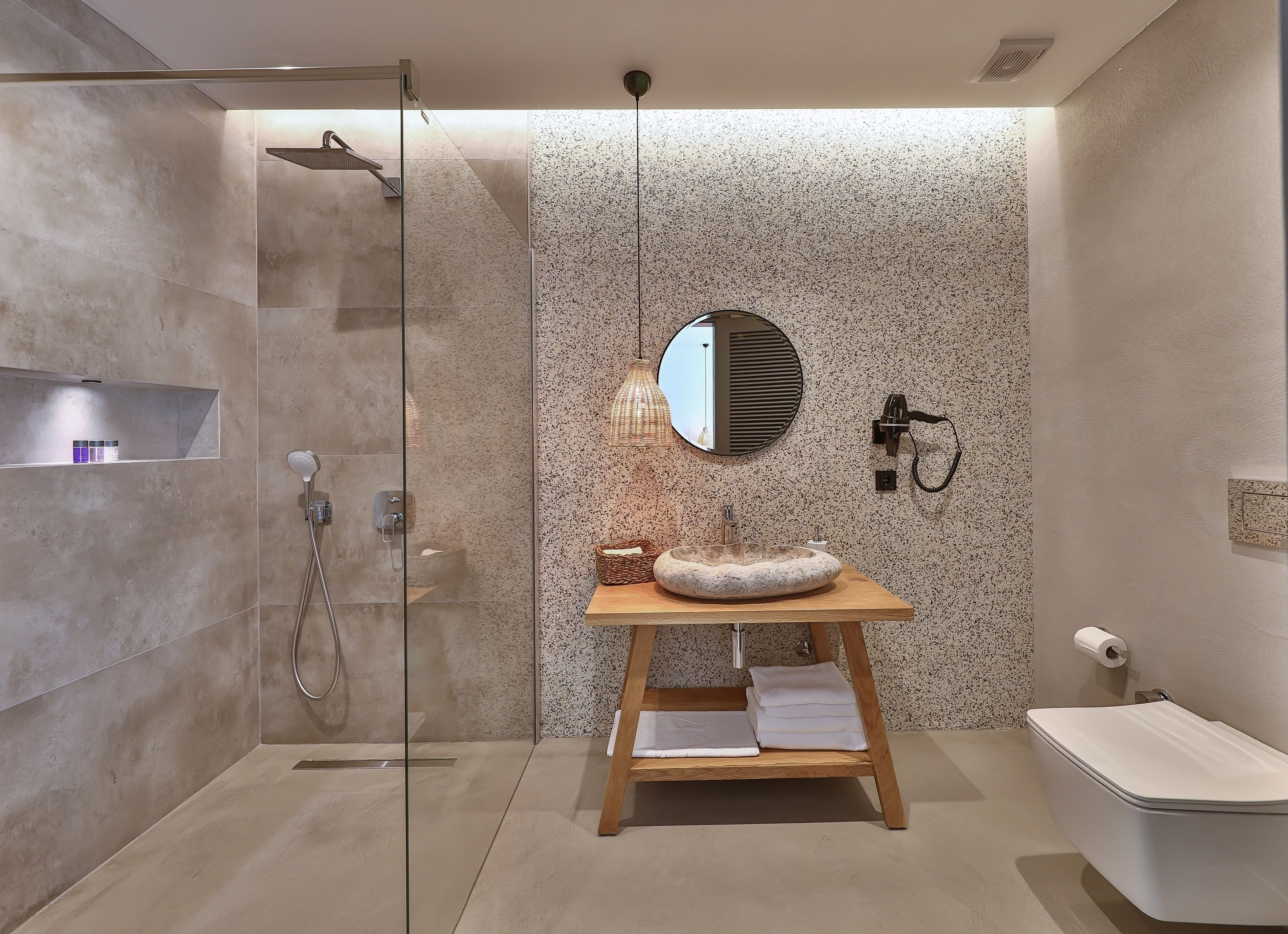 A modern bathroom with a glass shower enclosure, a round mirror, a wooden vanity with a stone sink, and a toilet.