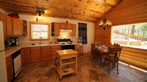 Interior de una cabaña de madera con cocina y comedor, que incluye armarios de madera, una estufa y una mesa de comedor con sillas.