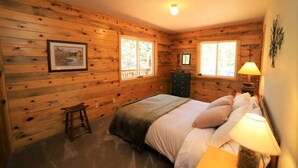 Un dormitorio de cabaña de madera con una cama, cómoda y dos ventanas.