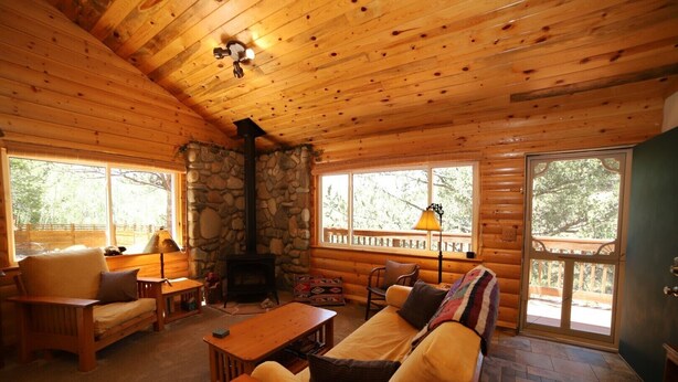 Interior acogedor de una cabaña con chimenea de piedra, muebles de madera y ventanas amplias con vistas al exterior.