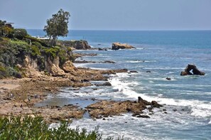 Litoral rochoso com ondas quebrando na praia, algumas pessoas caminhando pela areia e uma árvore no penhasco.