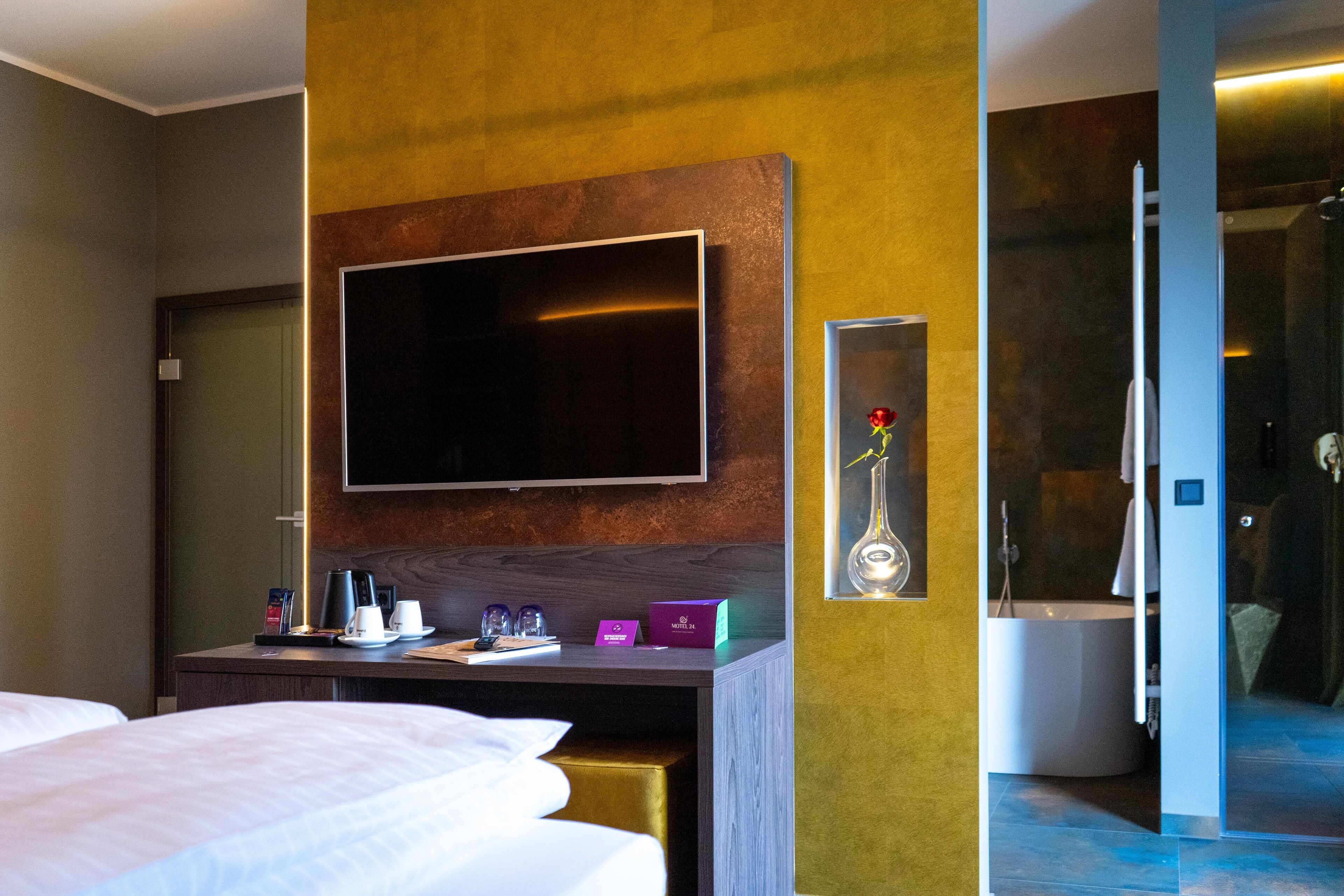 A hotel room with a bed, a desk with a flat-screen TV, a vase with a flower, and a bathtub.