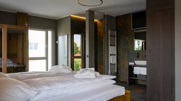 A modern hotel room with a large bed, a wooden bench, a bathroom with a sink and towel rack, and a view of the outdoors.