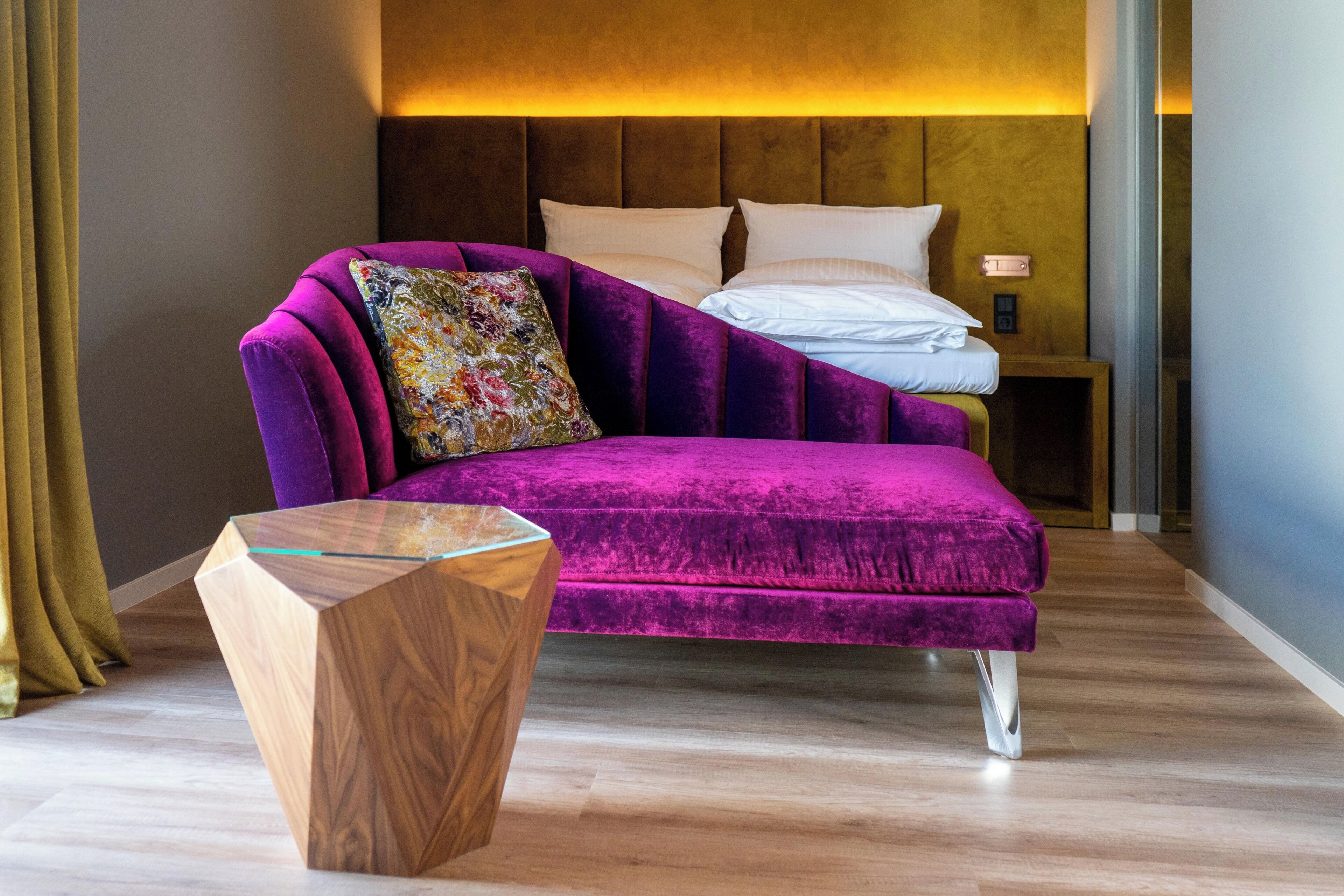 Una camera moderna con un divano in velluto viola, un tavolino in legno e un letto con lenzuola bianche.