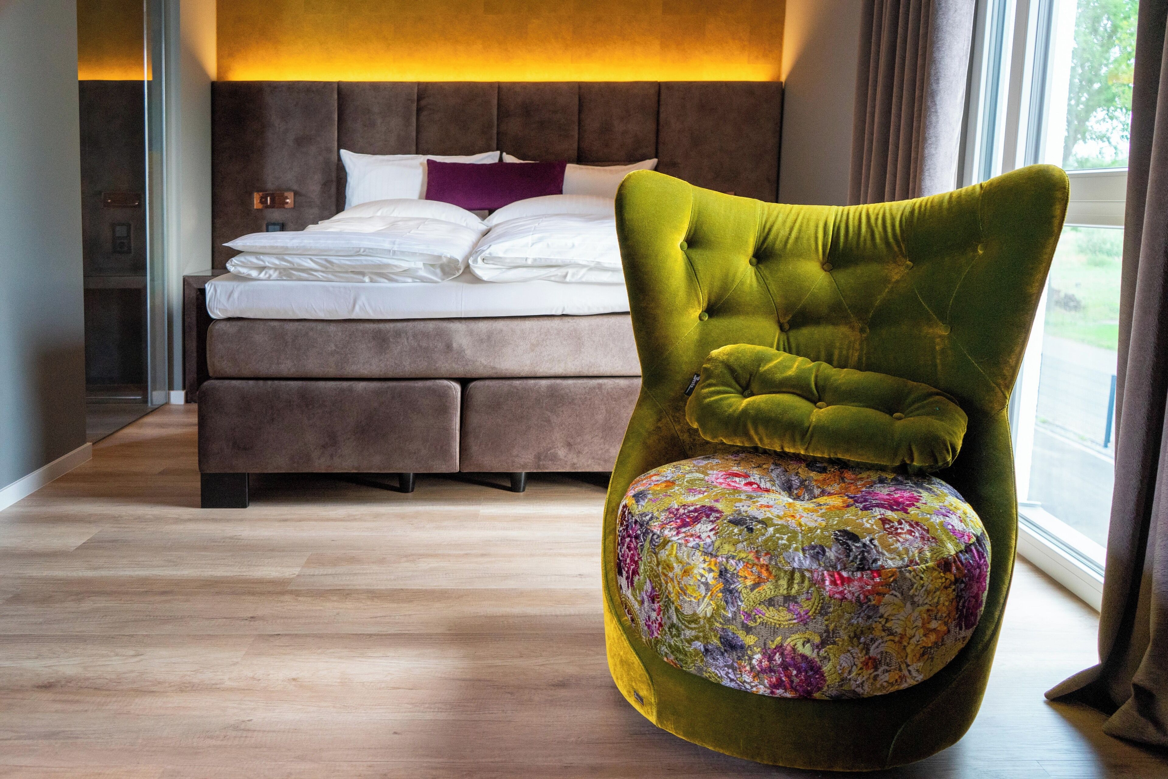A bedroom with a large bed, a green upholstered chair with a floral cushion, and a view of the outdoors through a window.