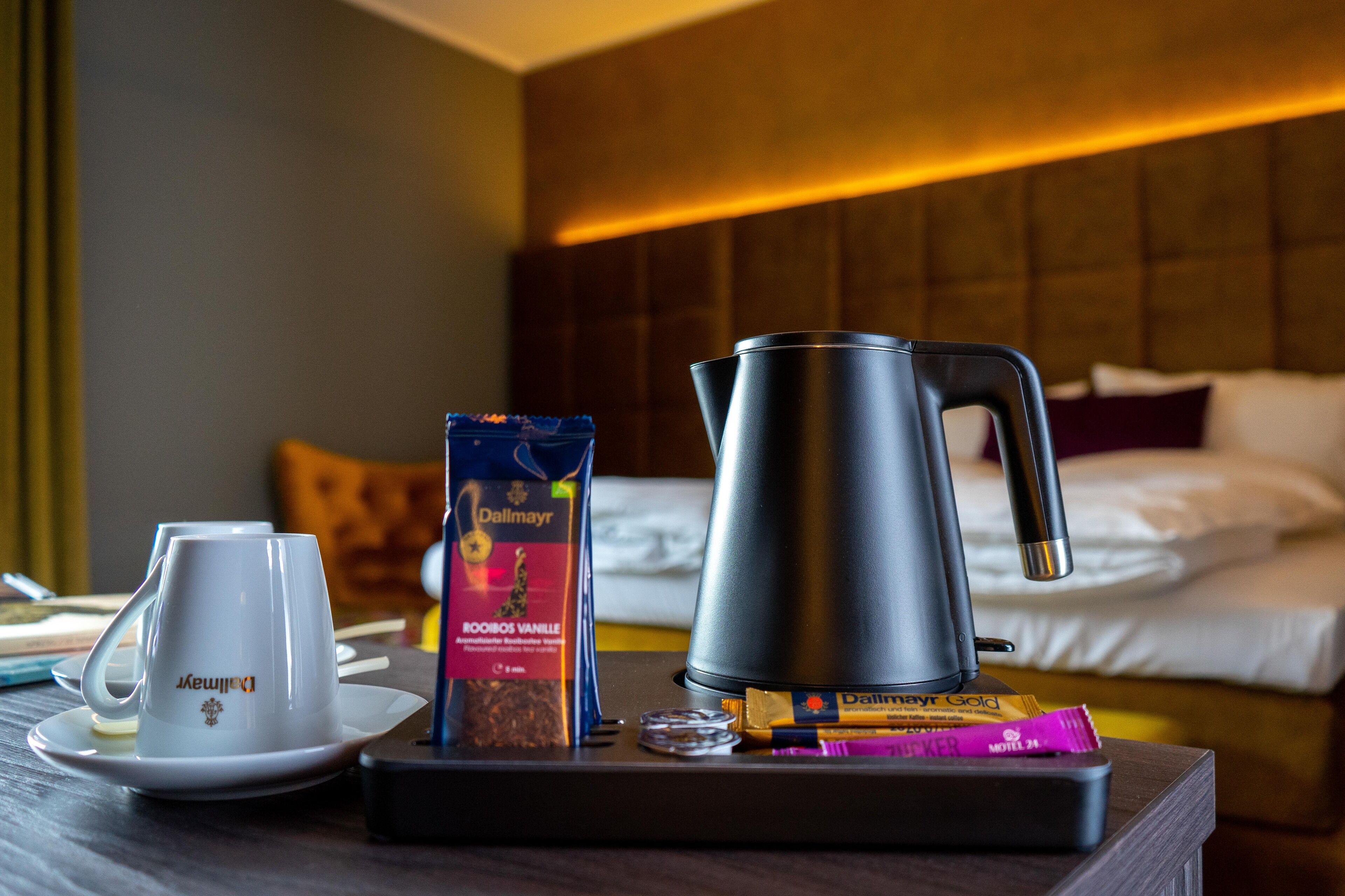 A hotel room with a bed, a bedside table with a coffee maker, a cup, and a box of chocolates.