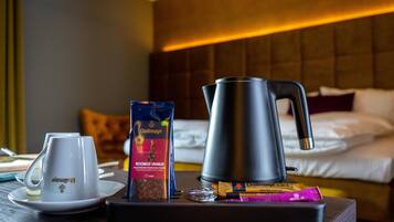 A hotel room with a bed, a bedside table with a coffee maker, a cup, and a box of chocolates.