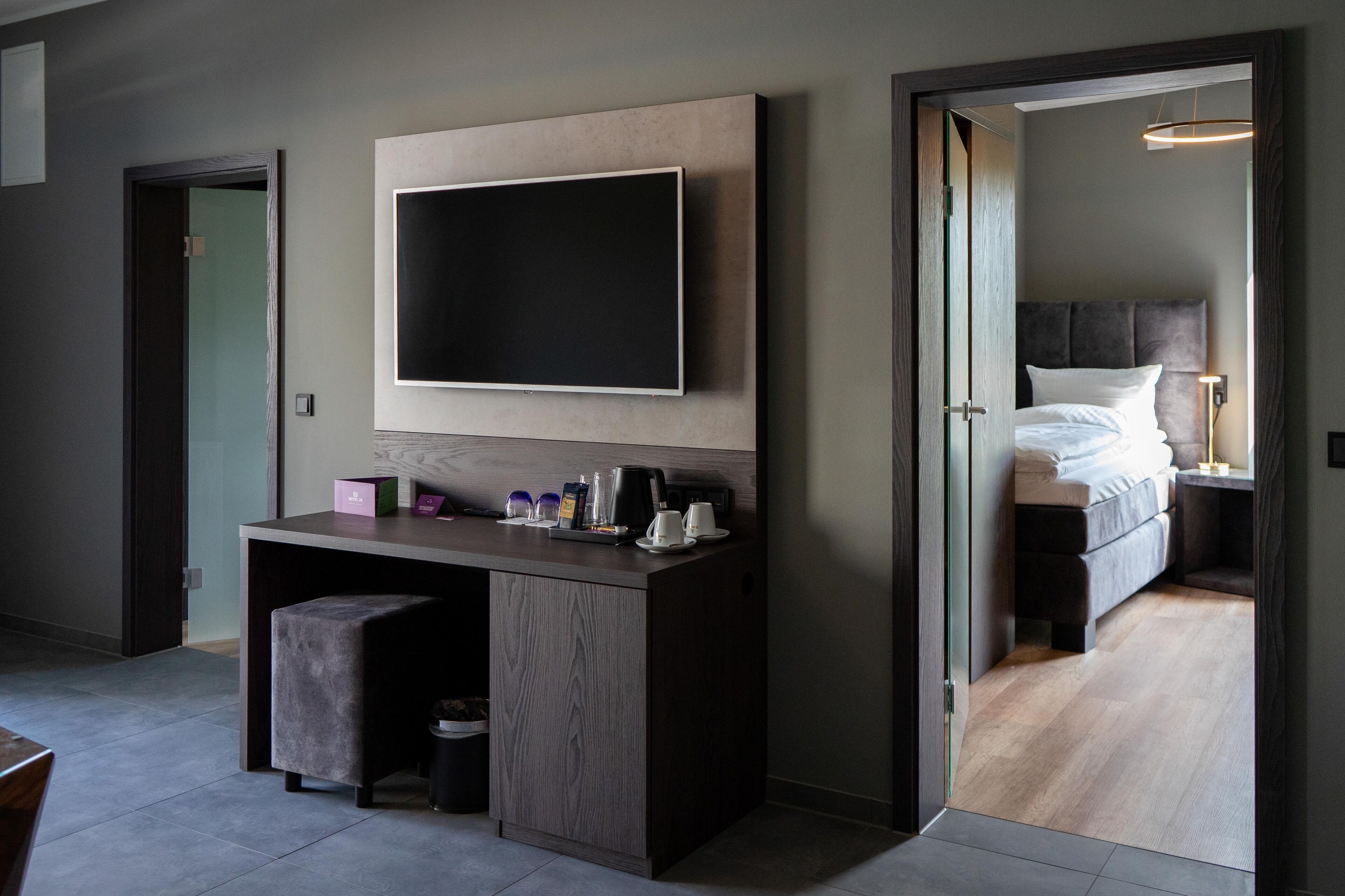 Una habitación de hotel moderna con un televisor de pantalla plana grande montado en la pared, un escritorio de madera con un taburete acolchado, una cama con cabecera oscura y una mesita de noche con una lámpara.