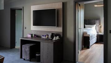 Una habitación de hotel moderna con un televisor de pantalla plana grande montado en la pared, un escritorio de madera con un taburete acolchado, una cama con cabecera oscura y una mesita de noche con una lámpara.