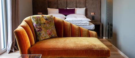 A modern hotel room with a plush orange sofa, a clear glass coffee table, and a neatly made bed with white linens and a purple accent pillow.