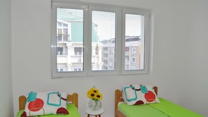 A room with two single beds, a small table with a vase of sunflowers, and a window with blinds.