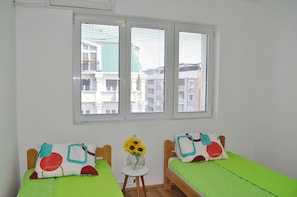 Two single beds with green bedspreads and abstract-patterned pillows, a small table with a vase of flowers, and a window with a city view.