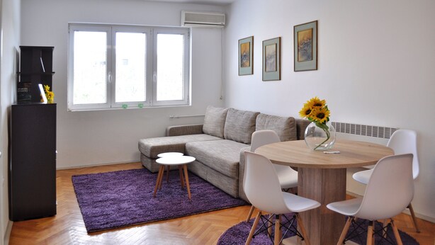A modern living room with a sofa, a dining table with chairs, a purple rug, and a vase with sunflowers.