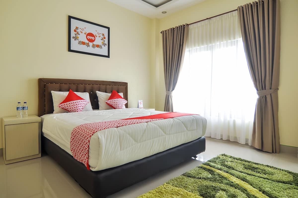 Een net opgemaakt bed met een deken in wit en rood, een bruin hoofdbord en twee kussens in rood en wit. Er staat een klein nachtkastje met twee waterflesjes en boven het bed hangt een prestatie in een lijst.
