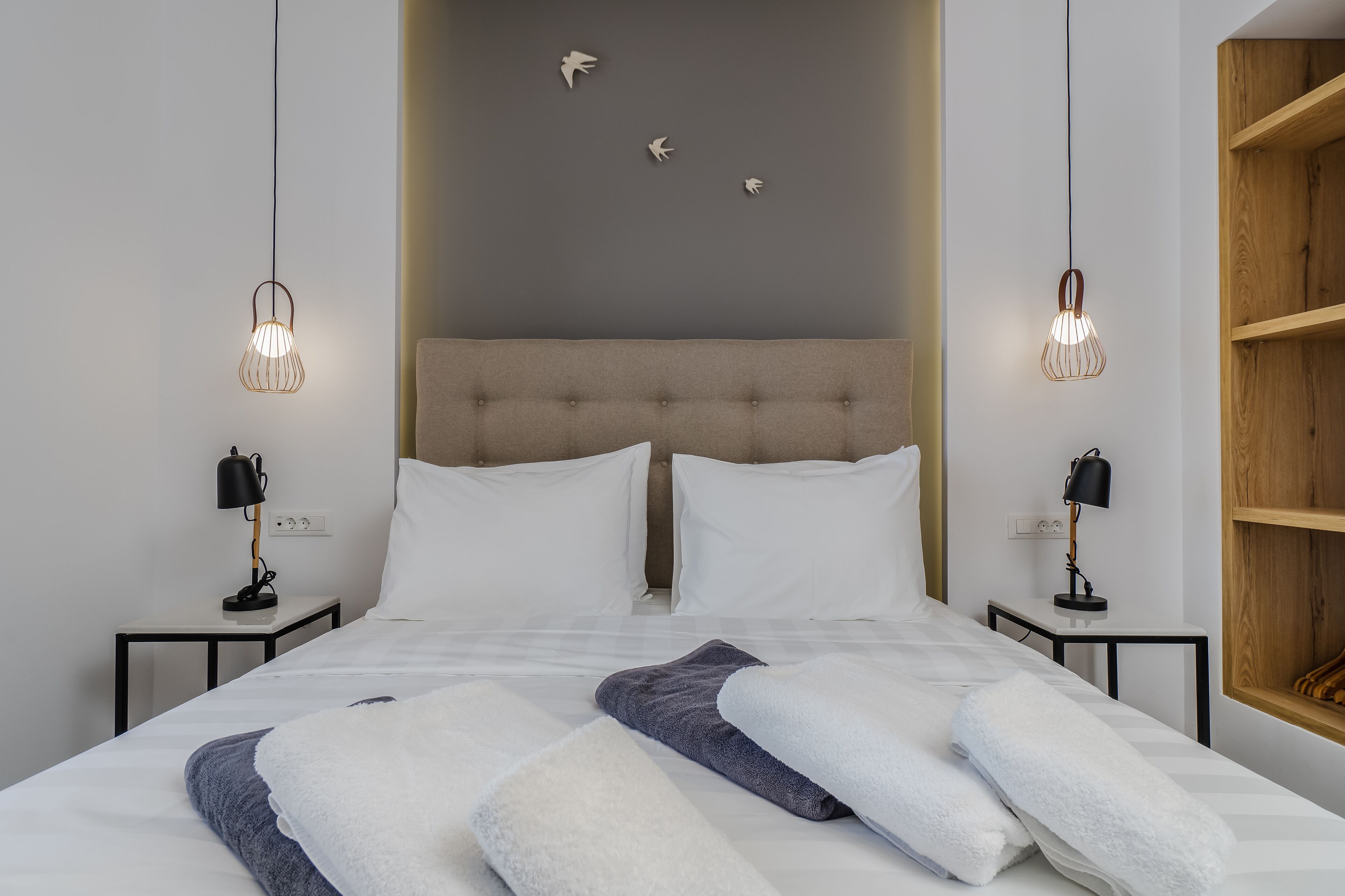 A neatly made bed with white and gray linens, two bedside tables with lamps, and a wooden shelf unit to the right.