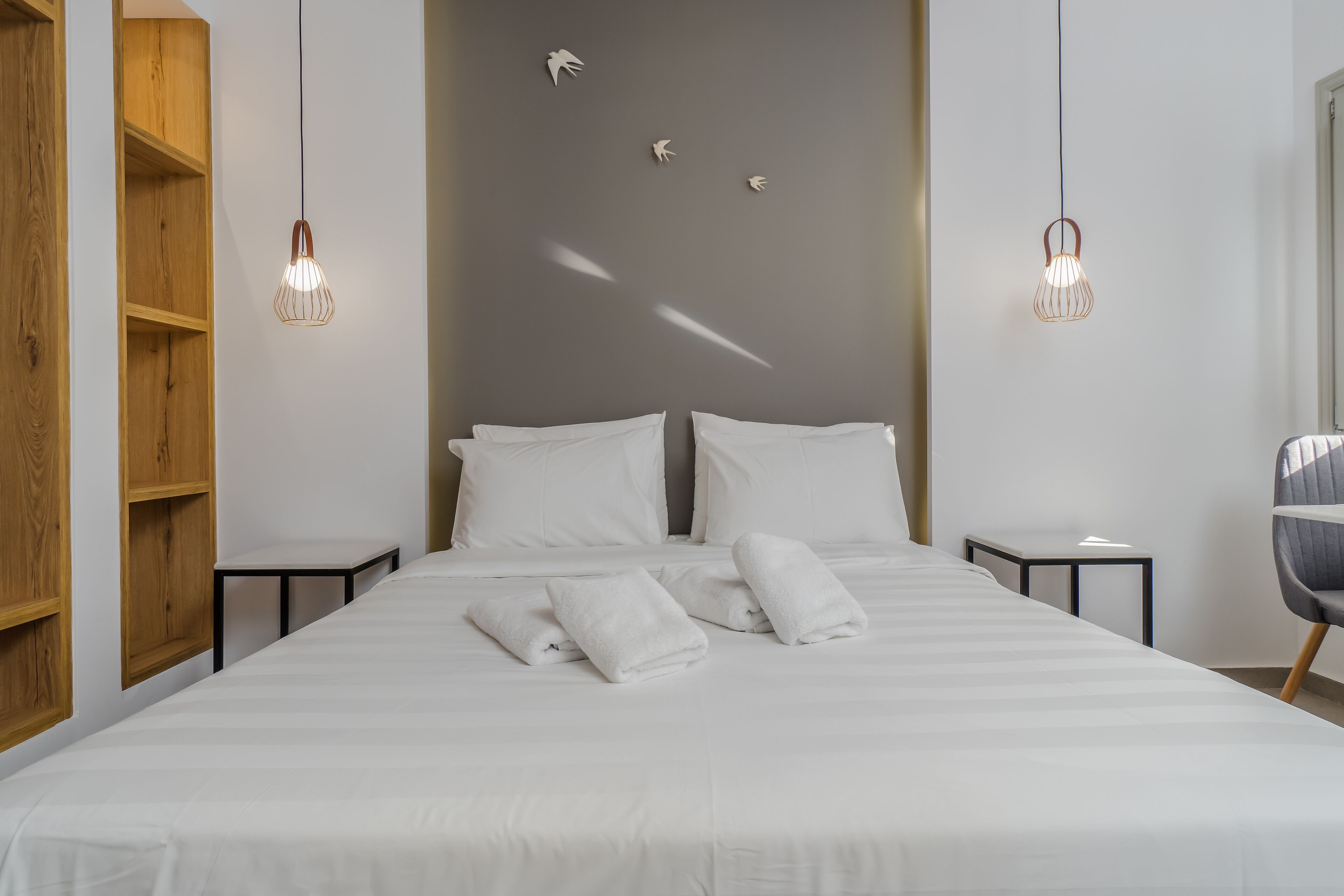 A neatly made bed with white linens and towels, two bedside tables, a wooden shelving unit, and two hanging pendant lights.