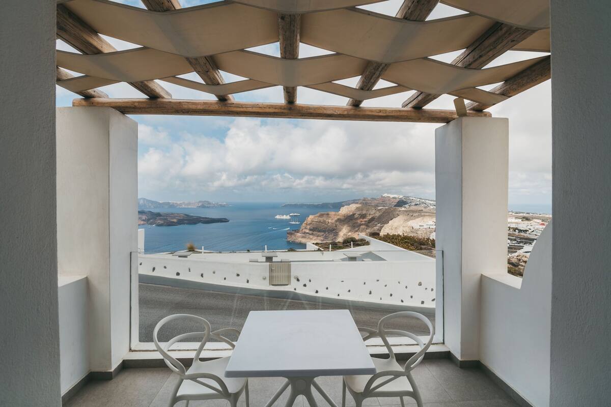 A balcony with a white table and chairs, offering a view of the sea and distant mountains.