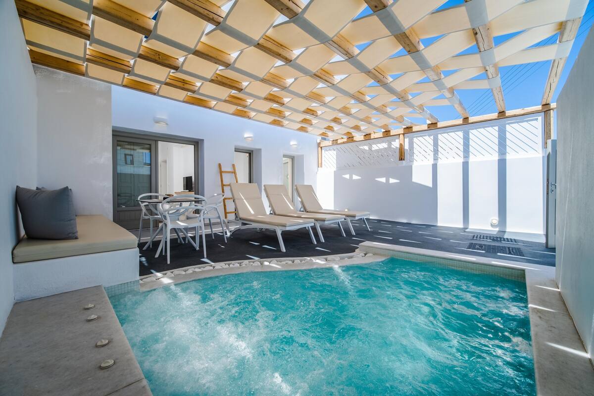 An indoor swimming pool with a wooden ceiling, lounge chairs, and a seating area with cushions.