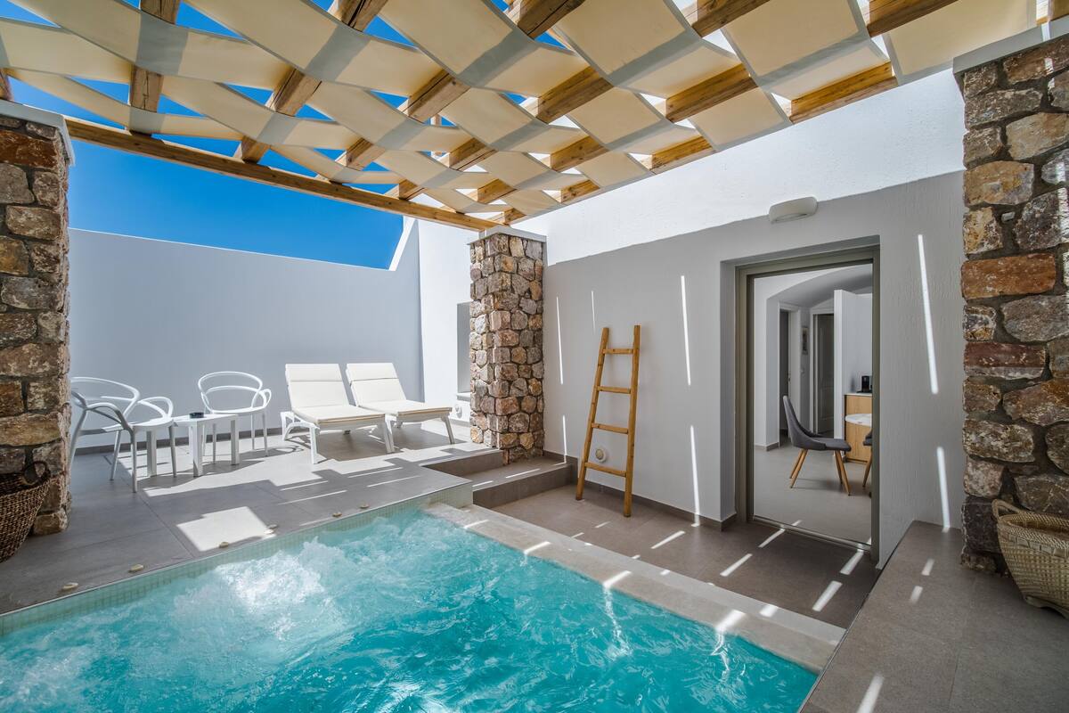 A pool area with a stone wall, wooden pergola, and white lounge chairs.