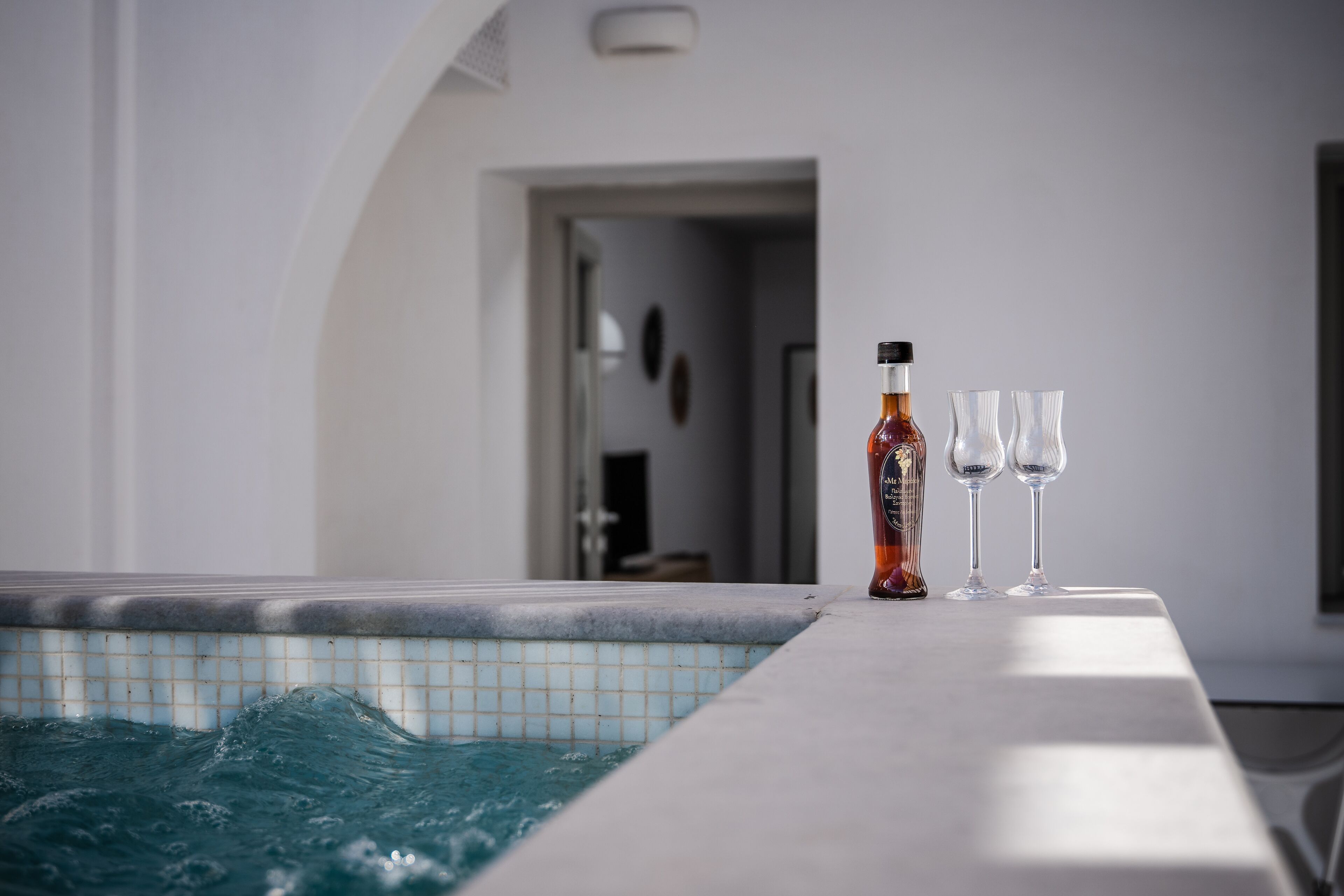 A poolside area with a tiled edge, a bottle, and two glasses on a table.