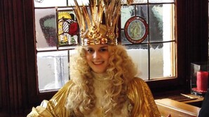 Una persona con túnica dorada y corona sentada a una mesa, con una ventana de vitrales al fondo.