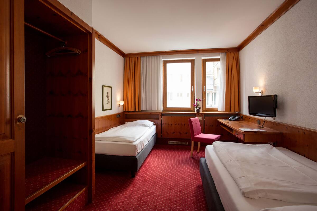 A hotel room with two beds, a desk with a chair, a TV, and a window with curtains.