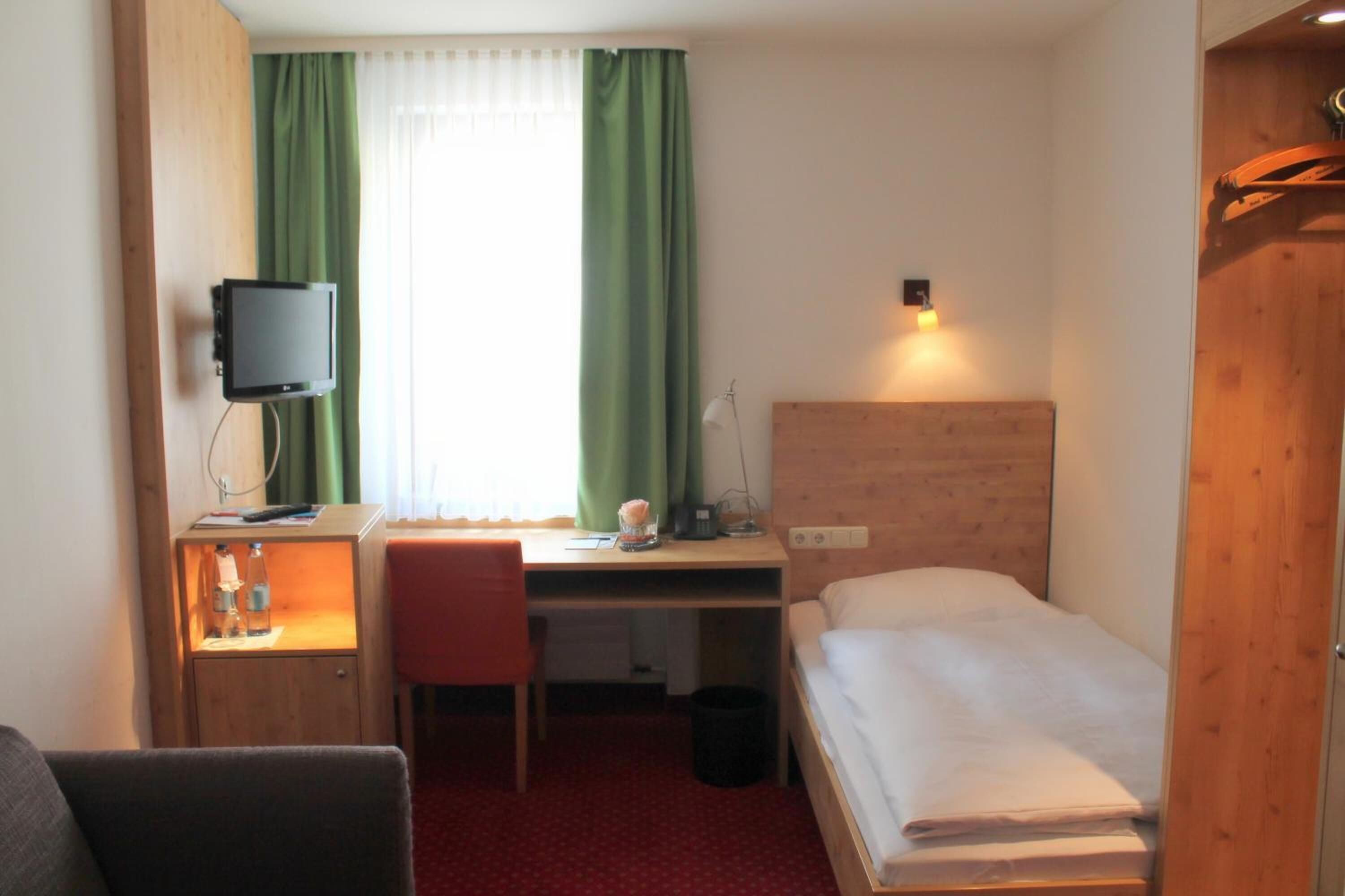 Hotellihuone, jossa on yksi sänky, työpöytä tuolilla, televisio ja ikkuna, jossa on vihreät verhot.