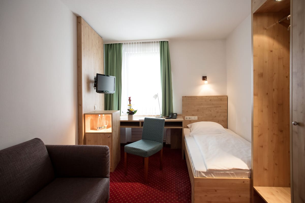 A compact hotel room with a wooden bed, a desk with a chair, a television mounted on the wall, and a small seating area with a sofa.