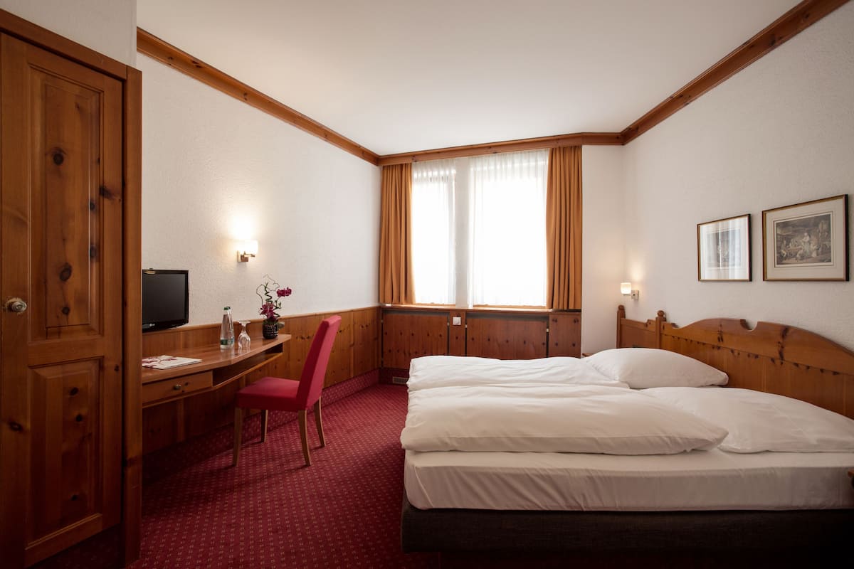 A hotel room with a wooden desk, a television, a red chair, a bed with white linens, and a window with curtains.