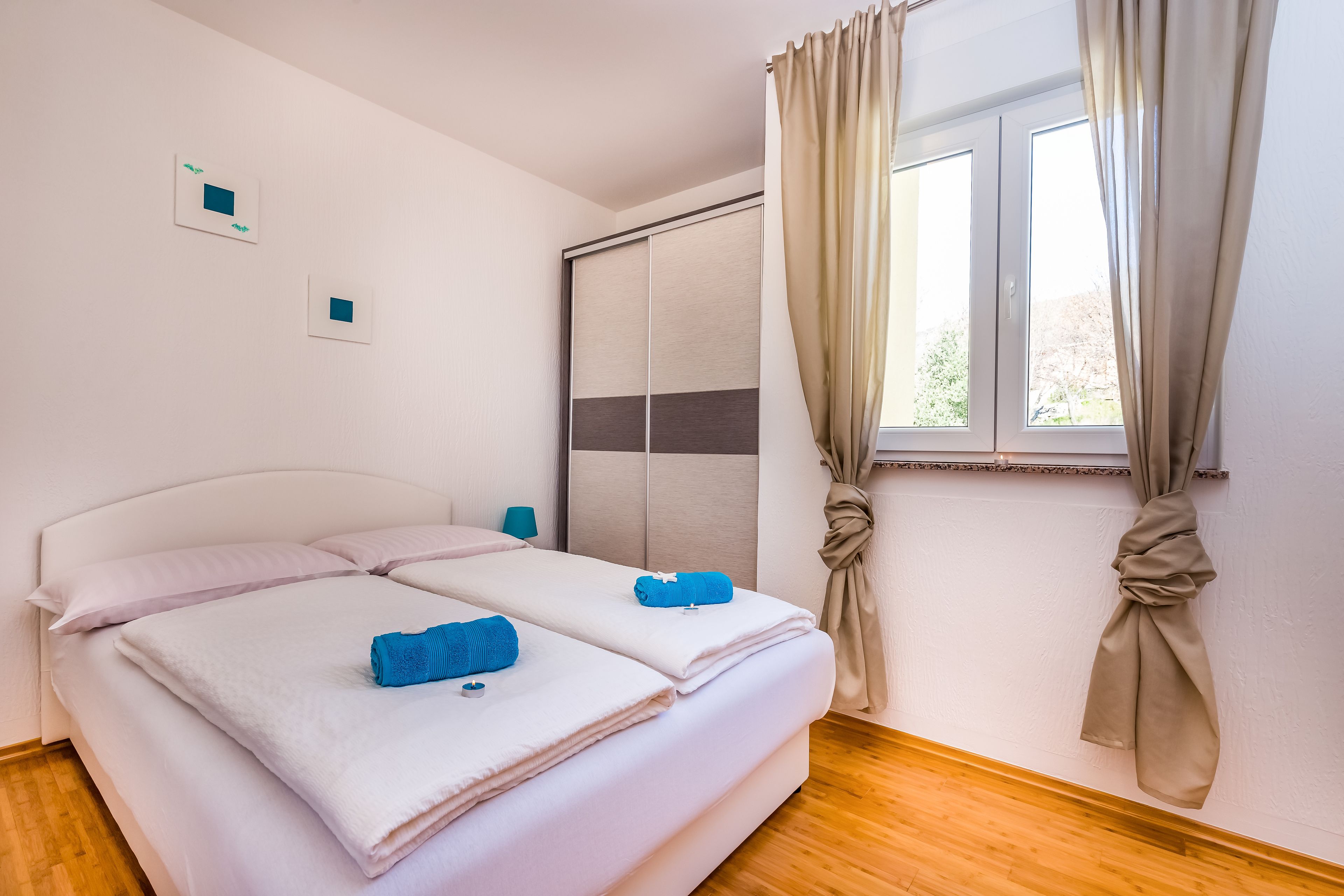 A double bed with white linens and blue towels, a sliding door closet, and a window with beige curtains.