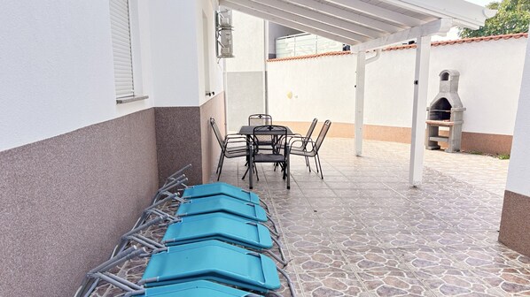 A covered outdoor area with a table and chairs, a barbecue grill, and a row of blue lounge chairs. - Villa Chiara (Crikvenica)