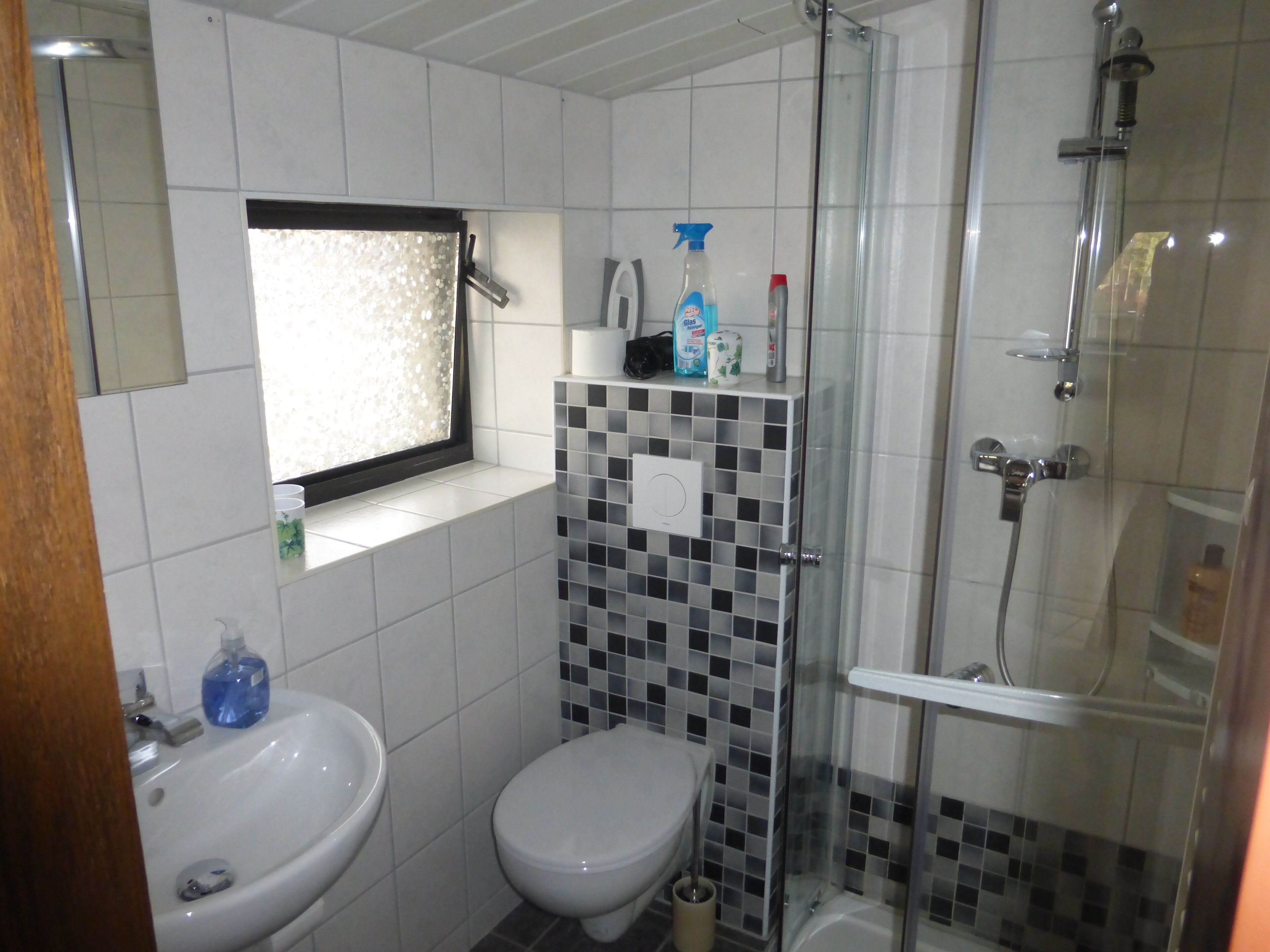 Une salle de bain avec un lavabo blanc, des toilettes, une douche avec une porte en verre et une étagère contenant des articles de toilette.