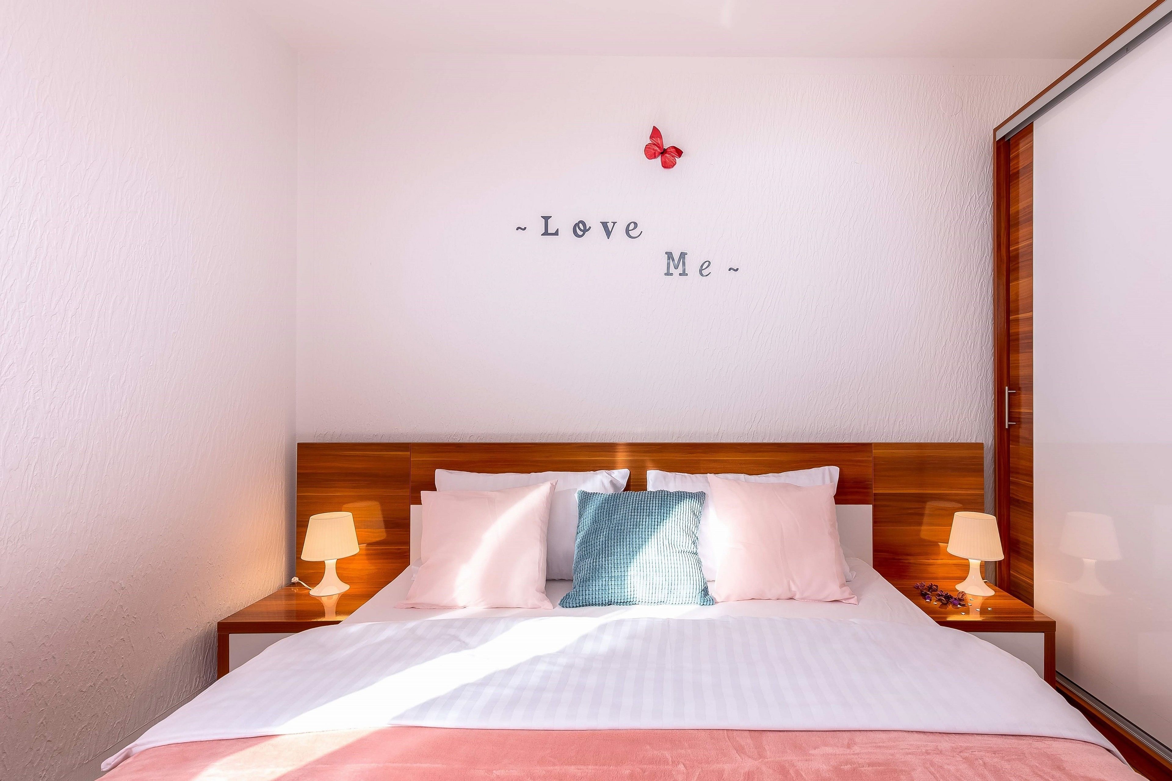 A bedroom with a bed, two bedside tables with lamps, a wall with a decorative phrase, and a wooden headboard.
