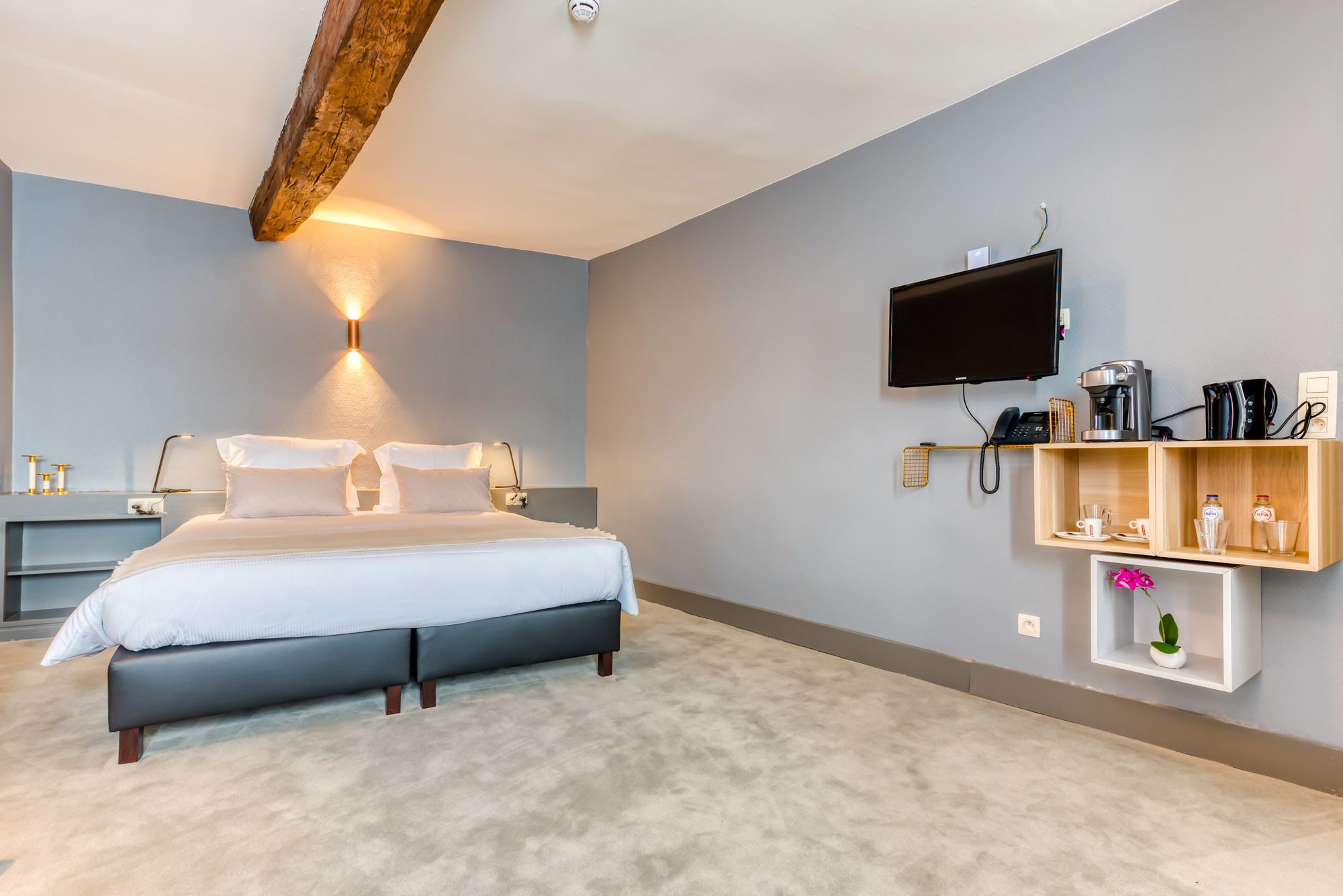 A modern hotel room with a large bed, a mounted TV, a coffee machine, and a shelf with toiletries.