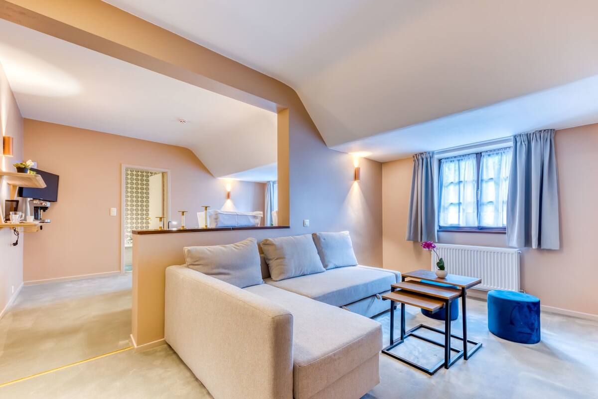 Un salon moderne avec un canapé, une petite table et un pouf bleu. Il y a une fenêtre avec des rideaux et un radiateur.