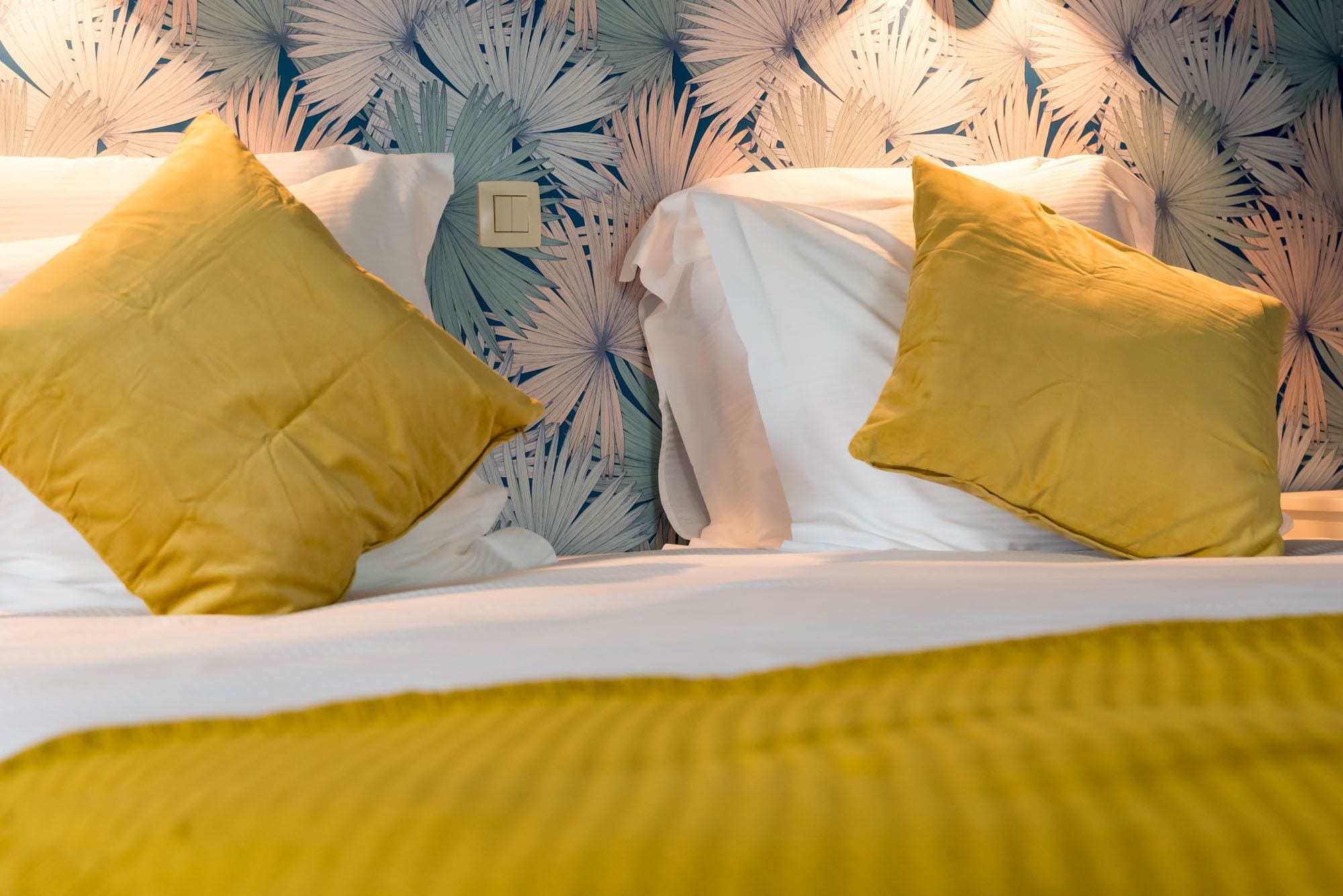 A bed with white linens and two yellow pillows, set against a wall with a tropical leaf pattern.