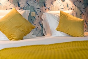 Una cama con sábanas blancas y dos almohadas amarillas, frente a una pared con estampado de hojas tropicales.