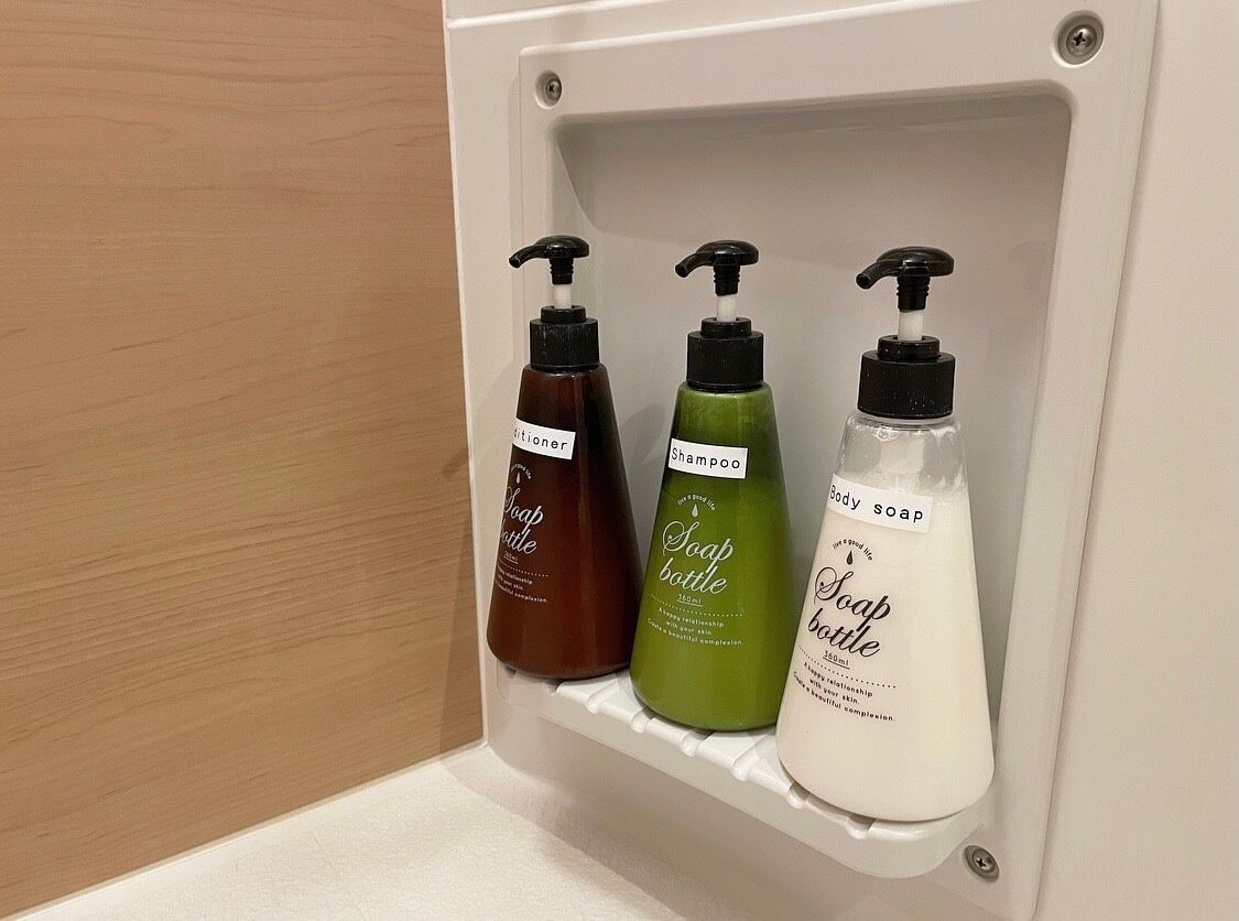Une étagère avec trois bouteilles de savon, étiquetées respectivement « Savon liquide », « Shampoing » et « Savon pour le corps ».