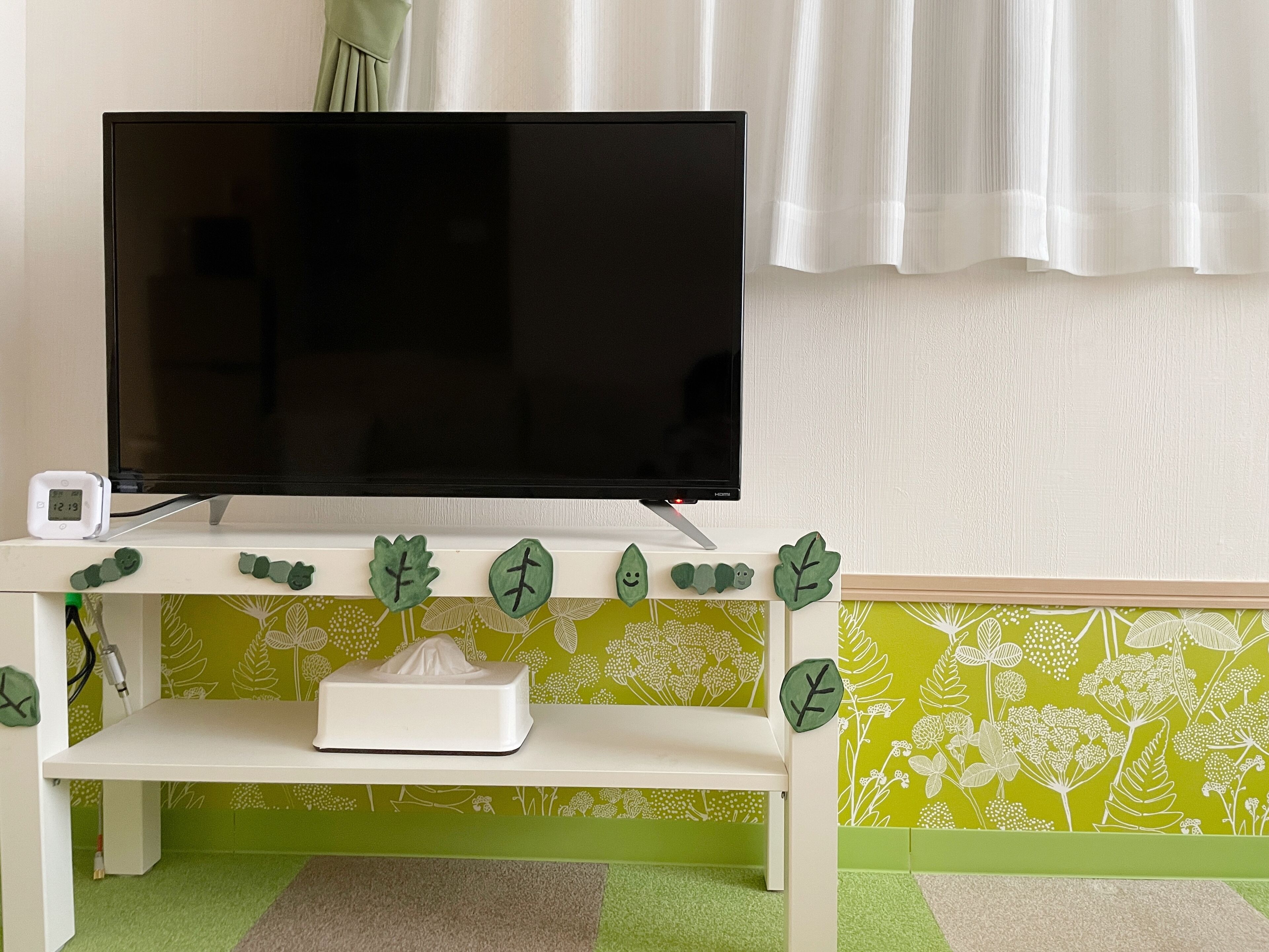 A television on a white stand with a green leaf decoration, a tissue box, and a clock on the left.