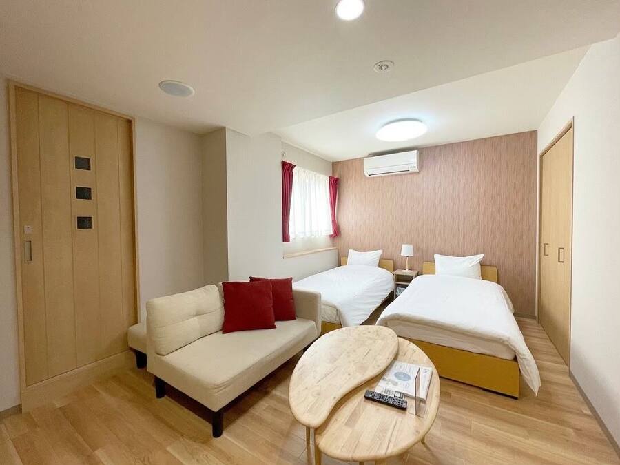 Quarto de hotel com sofá, duas camas de solteiro, uma mesa pequena, unidade de ar condicionado e guarda-roupa de madeira.