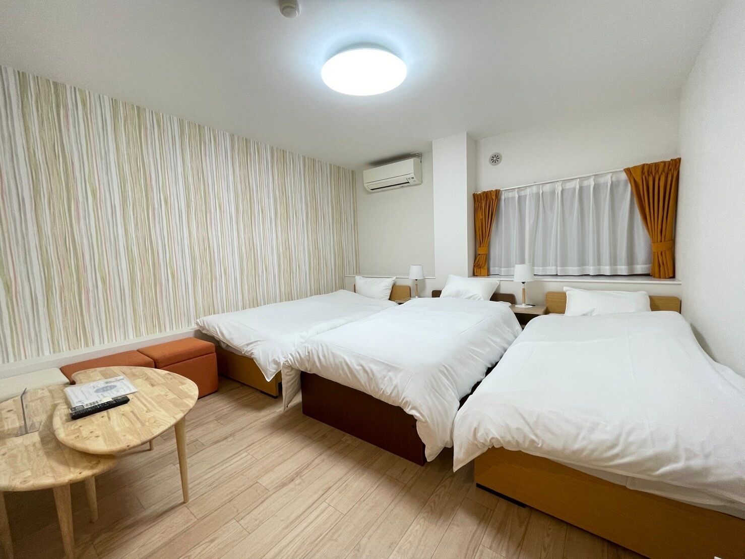 A hotel room with two beds, a small table, a sofa, a window with curtains, and an air conditioning unit.