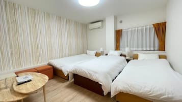 A hotel room with two beds, a small table, a sofa, a window with curtains, and an air conditioning unit.