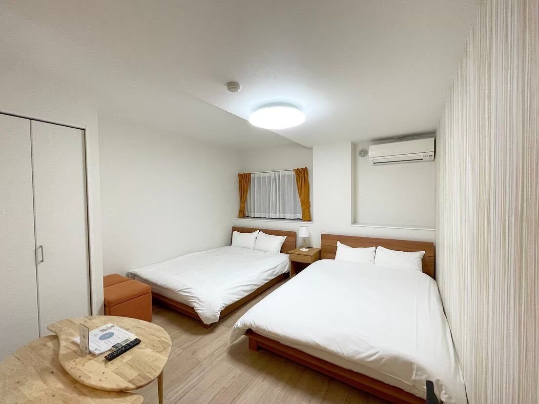 Hotellihuone, jossa on kaksi erillistä sänkyä, pieni puinen pöytä, yöpöytä lampulla, ikkuna verhoilla, ilmastointilaite ja kattovalaisin.