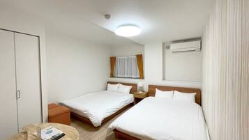 Hotellihuone, jossa on kaksi erillistä sänkyä, pieni puinen pöytä, yöpöytä lampulla, ikkuna verhoilla, ilmastointilaite ja kattovalaisin.