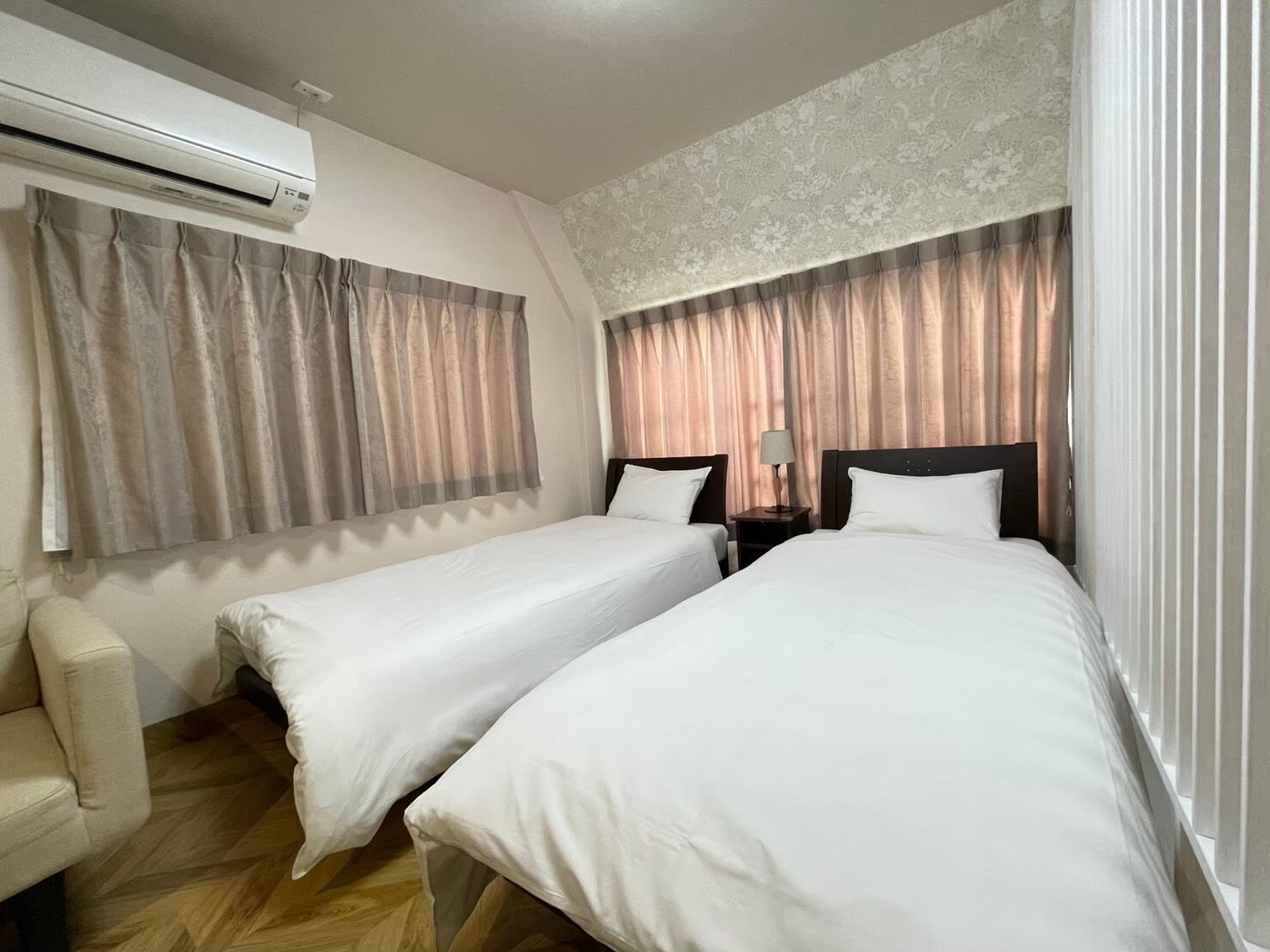 A hotel room with two single beds, a chair, an air conditioning unit, and a nightstand with a lamp.
