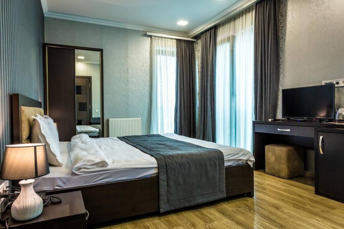 A hotel room with a bed, bedside table, lamp, television, and a view of the outside through large windows.