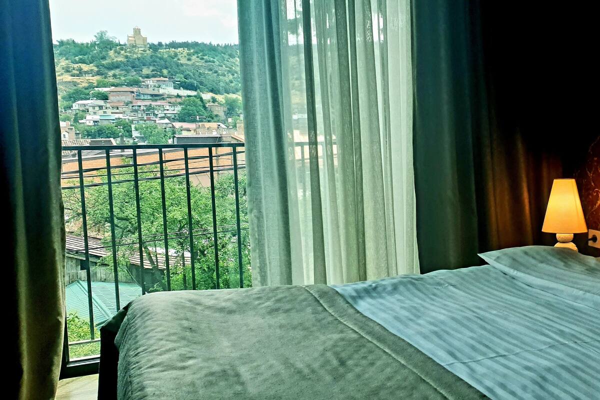 A hotel room with a bed, a nightstand with a lamp, and a view of a hillside town through the window.