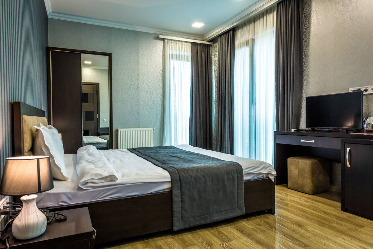 A hotel room with a bed, bedside table with a lamp, a desk with a television, and a large window with curtains.