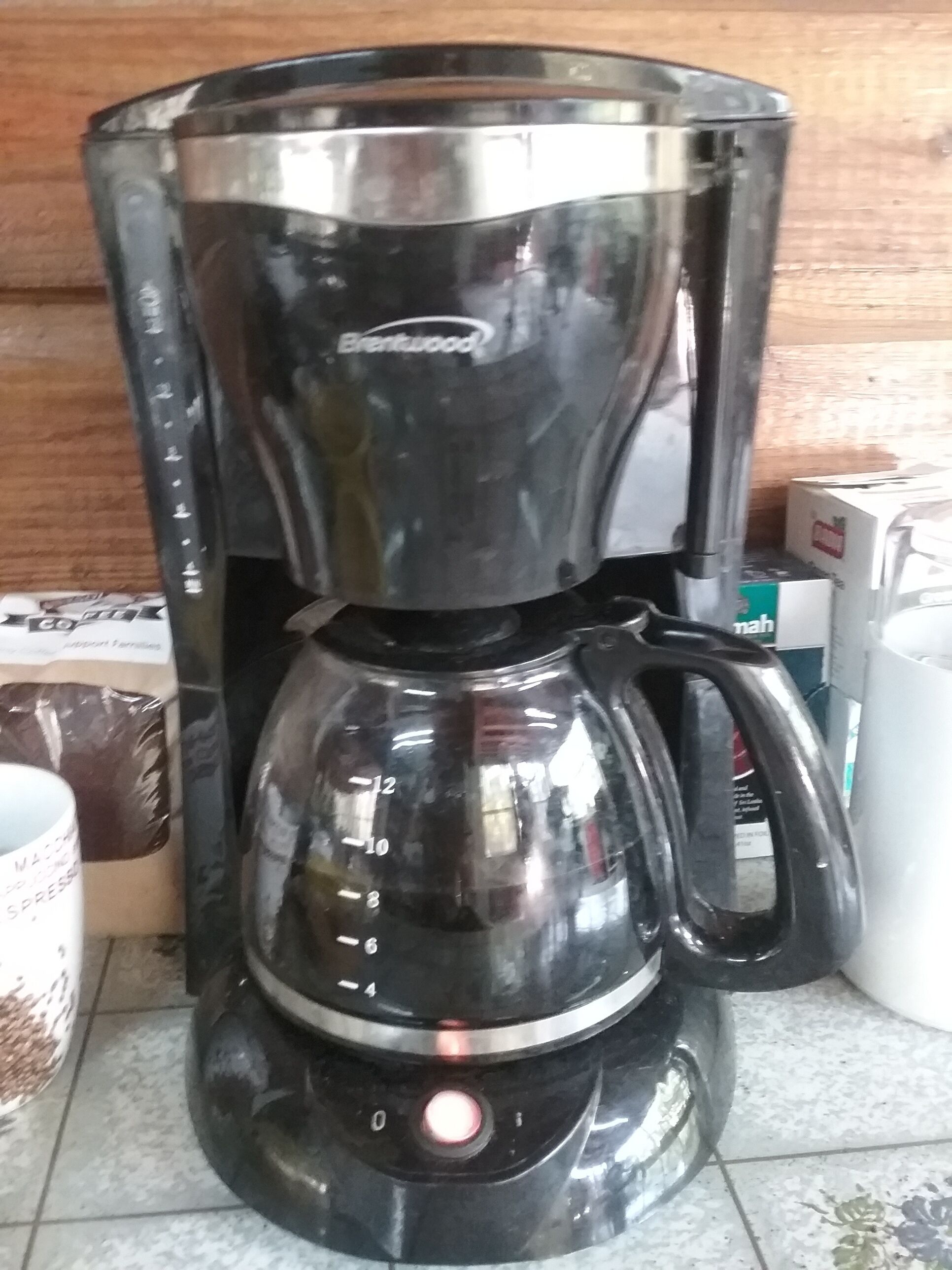 A black coffee maker with a glass carafe and a drip tray, placed on a tiled floor.