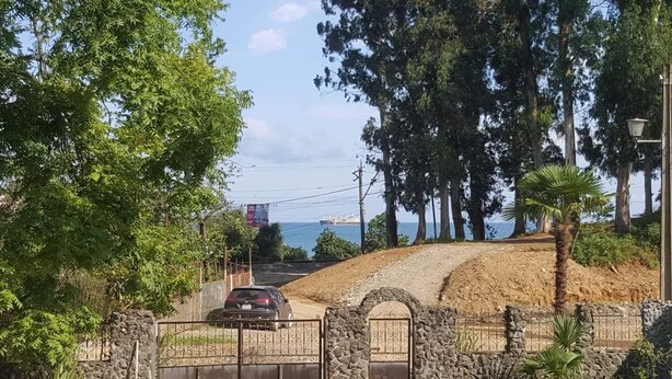 A coastal road with a car parked in front of a stone gate, leading to a sandy area with trees and a view of the sea. - beautiful sea garden (batumi)